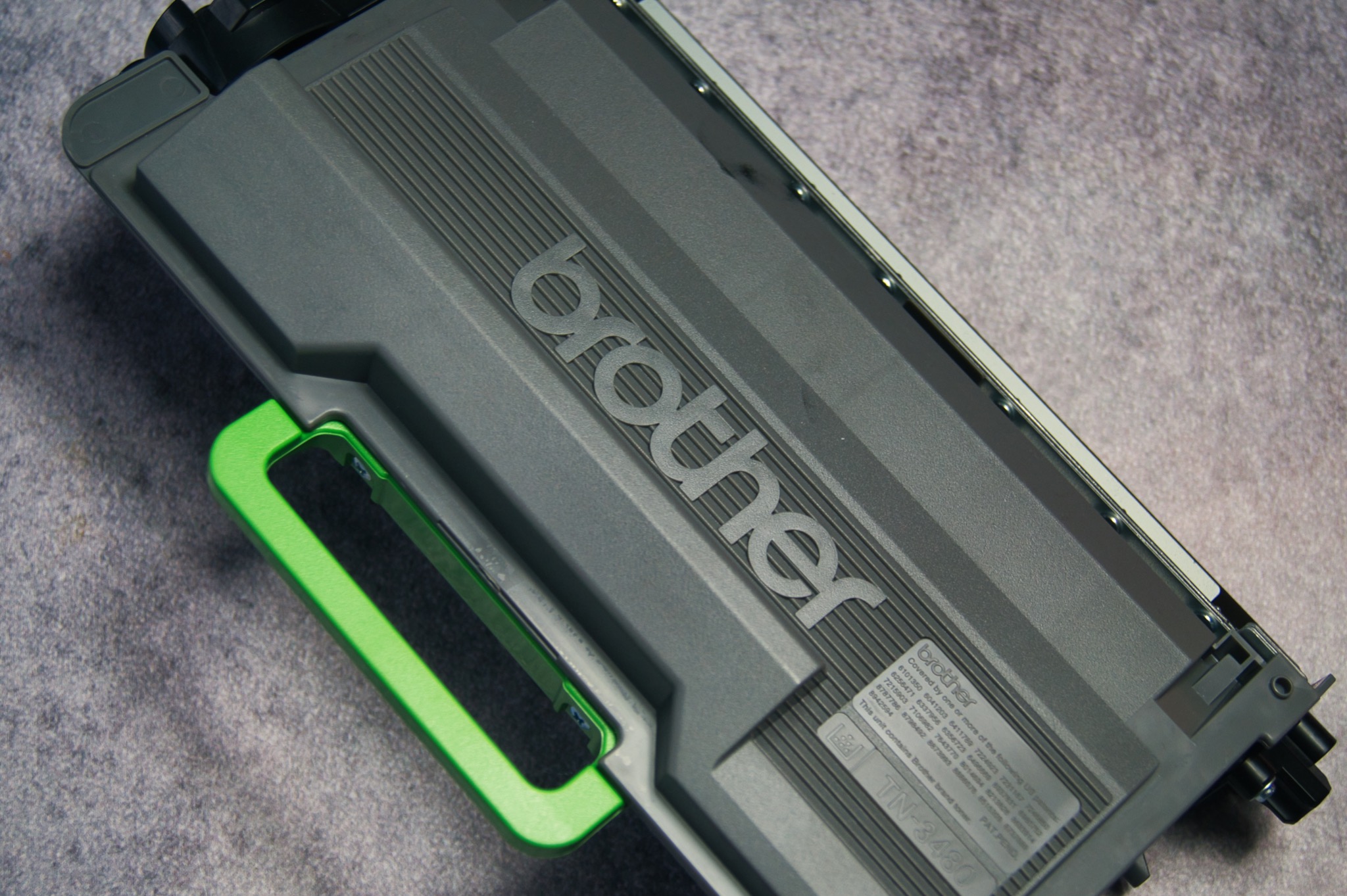 Szary toner do drukarki Brother z zielonym uchwytem, leżący na szarym tle.