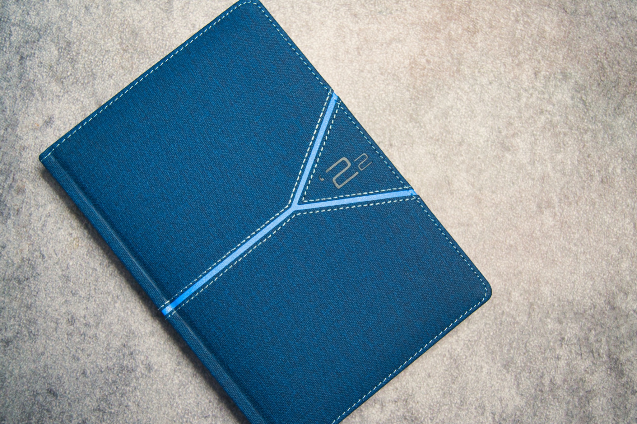 Zamknięty, granatowy notatnik z teksturowaną okładką, ozdobiony jasnoniebieskimi przeszyciami tworzącymi geometryczny wzór i subtelnym, srebrnym logo w prawym górnym rogu, leżący na szarym...