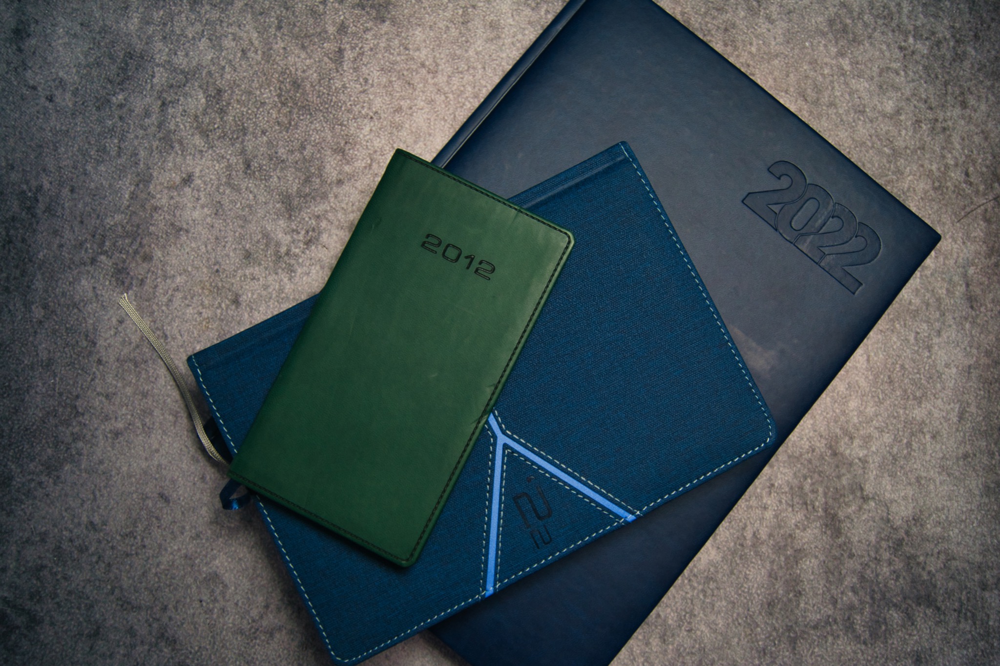 Stos trzech kalendarzy: ciemnozielony kalendarz z rokiem 2012 na wierzchu, niebieski kalendarz z geometrycznym wzorem i jasnoniebieskimi przeszyciami pośrodku oraz granatowy kalendarz z wytłoczonym...