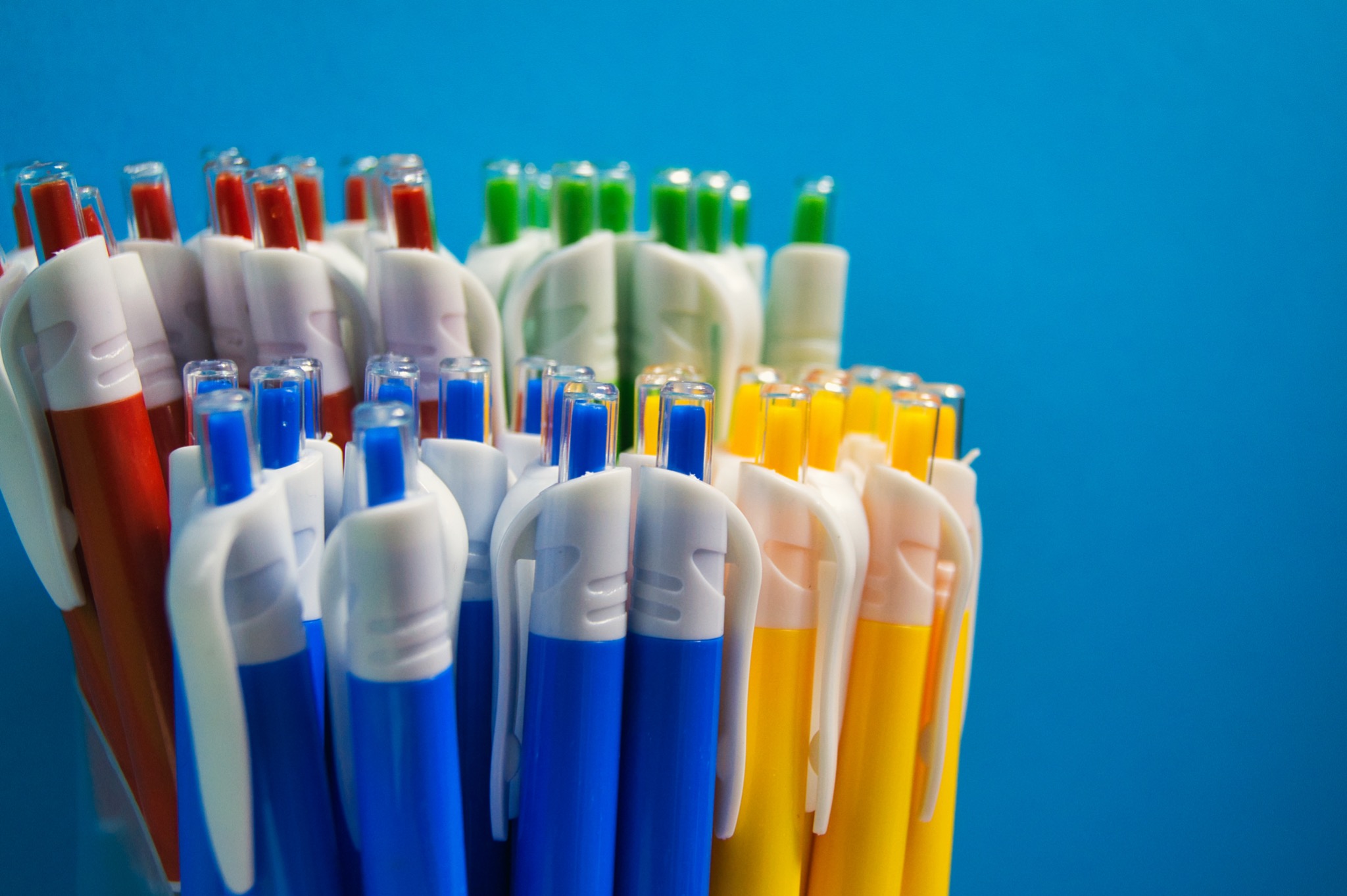Pęk kolorowych długopisów (niebieskie, żółte, czerwone, zielone) z białymi obudowami na jednolitym, jasnoniebieskim tle.