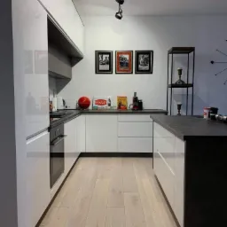 Nowoczesna kuchnia z białymi, lakierowanymi frontami, ciemnym blatem i drewnianą podłogą, oświetlona reflektorami sufitowymi, z dekoracjami w stylu retro.