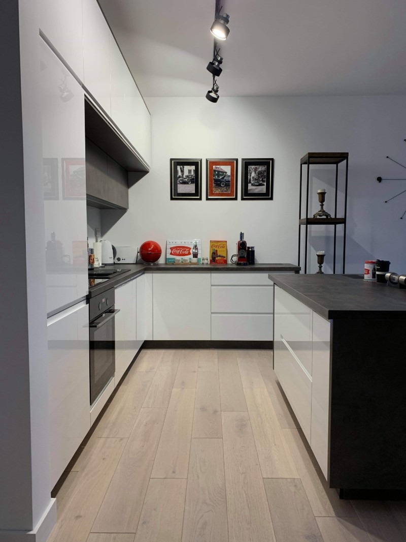 Nowoczesna kuchnia z białymi, lakierowanymi frontami, ciemnym blatem i drewnianą podłogą, oświetlona reflektorami sufitowymi, z dekoracjami w stylu retro.