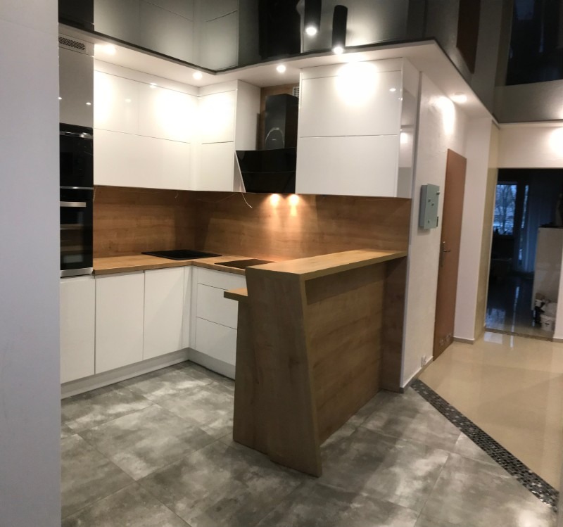 Nowoczesna kuchnia z białymi, lakierowanymi frontami, drewnianym blatem i barkiem, czarnym okapem oraz błyszczącym sufitem podwieszanym.