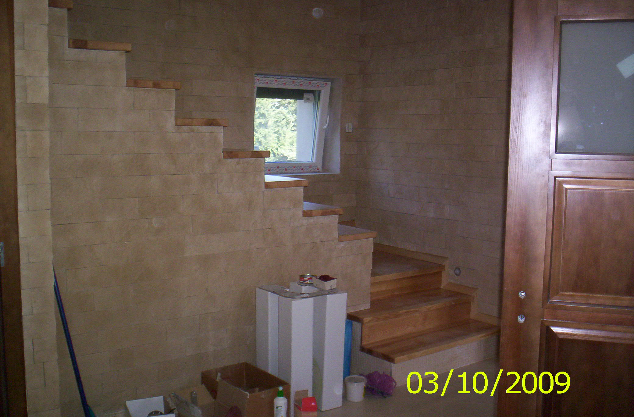 Wykończone schody z drewnianymi stopniami i okładziną ścienną imitującą kamień, widoczne narzędzia i materiały budowlane w tle, data wykonania zdjęcia (03/10/2009) w prawym dolnym rogu.