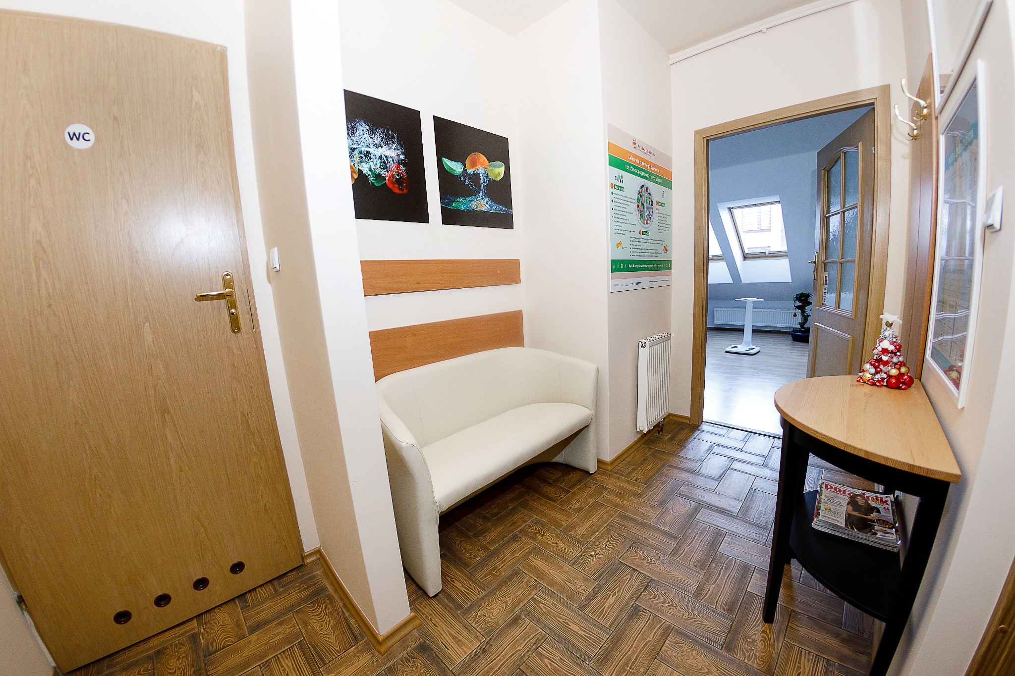 Wnętrze gabinetu: korytarz z białą kanapą, plakatami z owocami i warzywami, stolikiem z dekoracją świąteczną, oraz wagą diagnostyczną widoczną w głębi pomieszczenia.