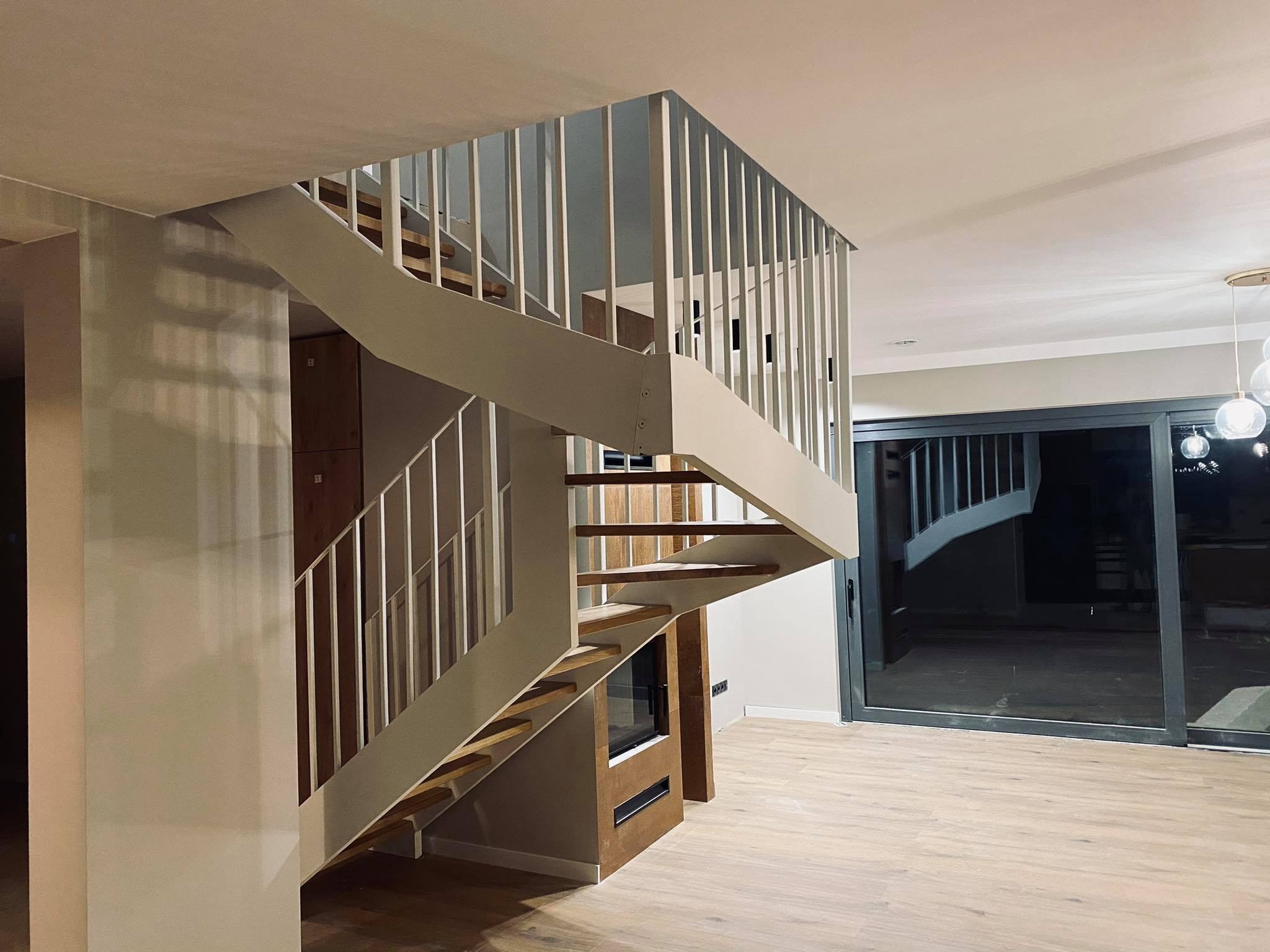 Nowoczesne schody drewniane z białymi barierkami w jasnym wnętrzu domu, zintegrowane z zabudową meblową i kominkiem pod biegiem schodów, widok na przesuwne drzwi tarasowe.