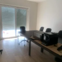 Jasne biuro z dużym oknem z żaluzjami, ciemnym stołem konferencyjnym, krzesłami obrotowymi i ekspresem do kawy.