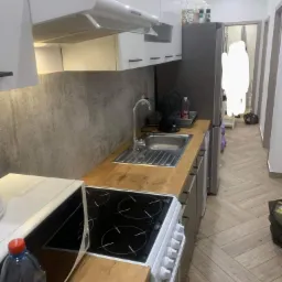 Kompaktowa kuchnia z drewnianym blatem, widok na zlew, płytę grzewczą i białe szafki, butelka wody mineralnej w pierwszym planie, czarny wentylator obok zlewu, w tle widoczny korytarz.