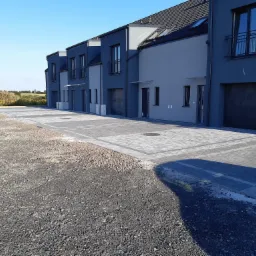 Nowoczesne szeregówki z szarego betonu z wybrukowanym podjazdem z kostki w dwóch odcieniach szarości, teren przed budynkami jeszcze niezagospodarowany.