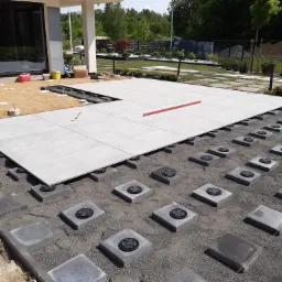 Fragment tarasu w trakcie budowy: szare płyty tarasowe ułożone na regulowanych wspornikach, widoczne przygotowane podłoże z kostki brukowej, poziomica na płytach, dom w tle.