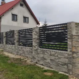 Nowoczesne ogrodzenie panelowe z poziomymi, czarnymi przęsłami i betonowymi słupkami o nieregularnej fakturze, na tle domu z czerwoną dachówką i pochmurnego nieba.