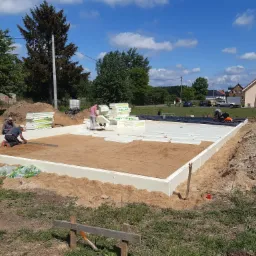 Budowa fundamentów: robotnicy układają białe płyty izolacyjne na przygotowanym podłożu, w tle widoczny dom jednorodzinny i stosy materiałów budowlanych.