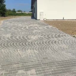 Długa, szara, prostokątna nawierzchnia z kostki prowadząca wzdłuż białego budynku z ciemnymi oknami; świeżo ułożona kostka zasypana piaskiem, teren wokół jeszcze niezagospodarowany.