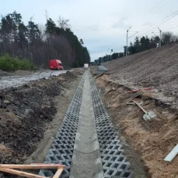 Koryto odwadniające z betonowych ażurowych płyt na placu budowy, widoczna czerwona furgonetka w tle, ziemia, drzewa i słupy trakcji elektrycznej.