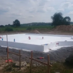 Białe, zaizolowane fundamenty domu z wystającymi rurami, otoczone ziemią i drewnianymi stemplami, w tle zielony krajobraz i pochmurne niebo.