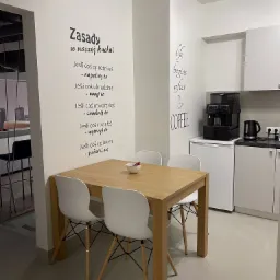Jasna kuchnia biurowa z drewnianym stołem otoczonym białymi krzesłami, na ścianie naklejka z zasadami korzystania z kuchni, ekspres do kawy i czajnik na blacie.