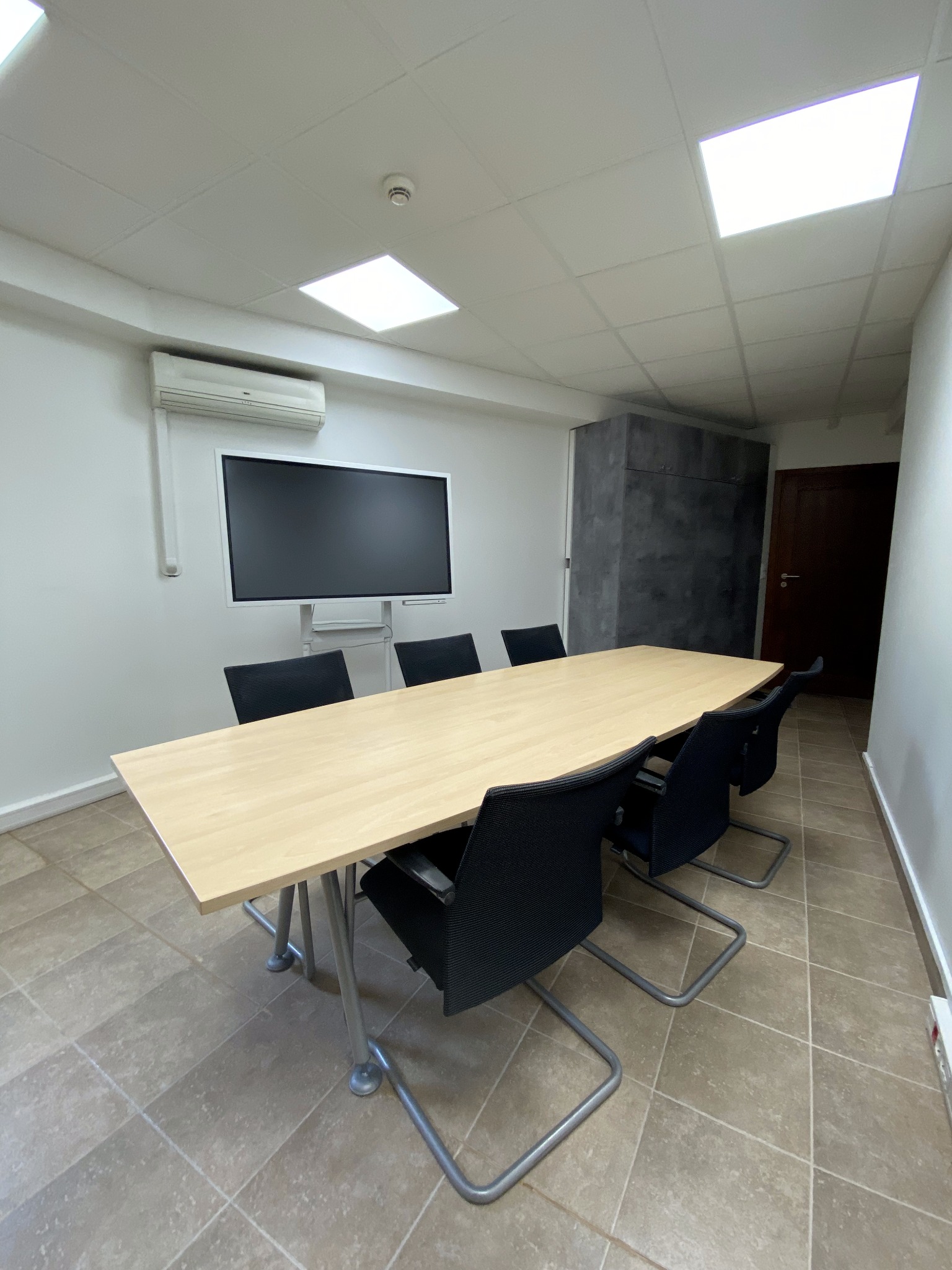 Sala konferencyjna z długim stołem, sześcioma krzesłami, ekranem na ścianie i klimatyzacją, oświetlona panelami LED w suficie.