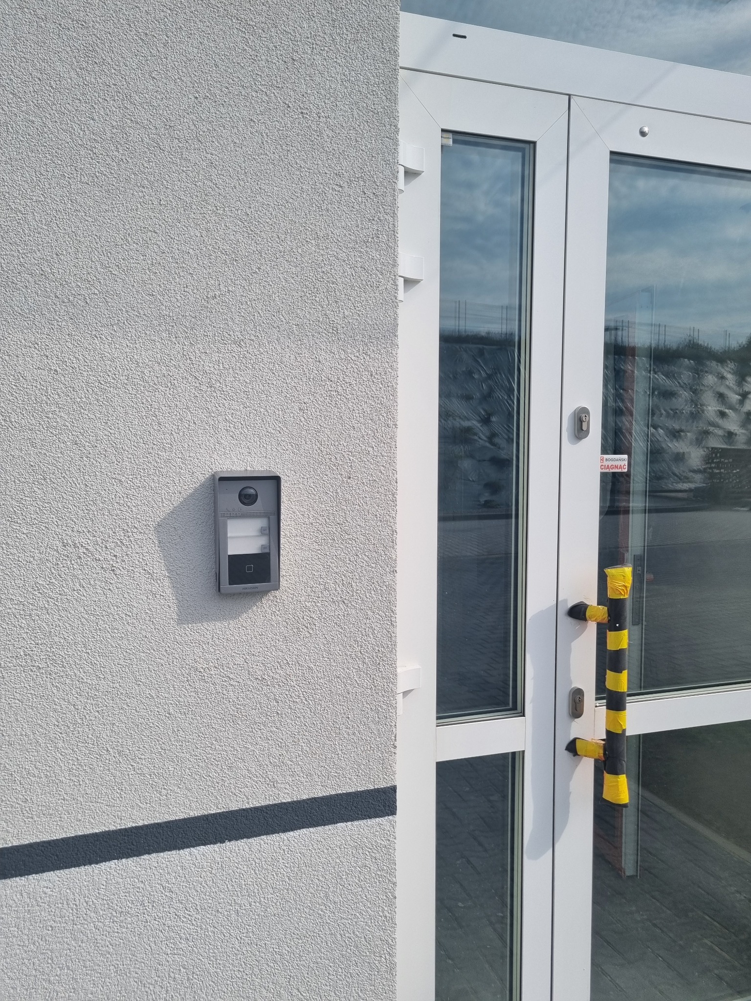 Szary wideodomofon Hikvision z kamerą, przy wejściu do budynku z białymi drzwiami i czarno-żółtymi zabezpieczeniami na klamce.