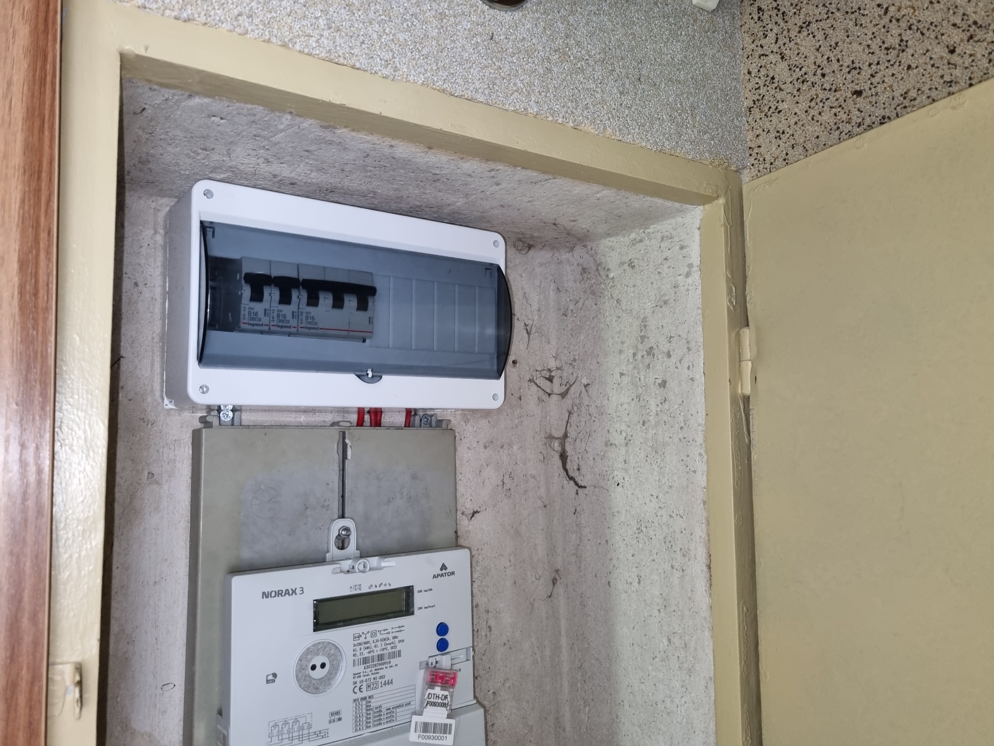 Szara skrzynka bezpiecznikowa i licznik energii elektrycznej Norax 3 na ścianie w Starej Wsi, widoczne ślady użytkowania i niedoskonałości wykończenia.