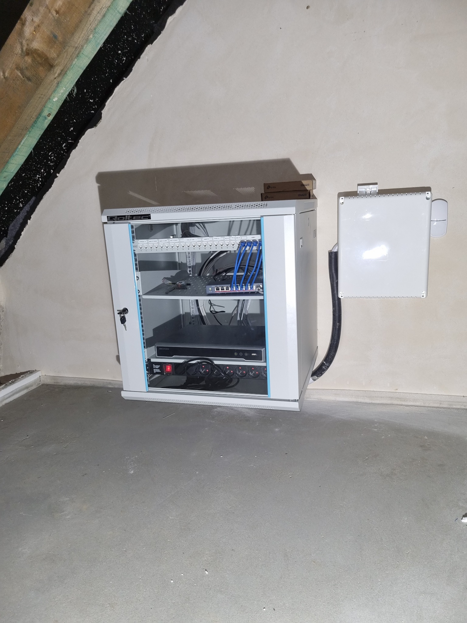 Szafa rackowa z urządzeniami sieciowymi i rejestratorem, zabezpieczona kluczem, obok puszka instalacyjna z kablem w peszlu, na tle surowego poddasza.
