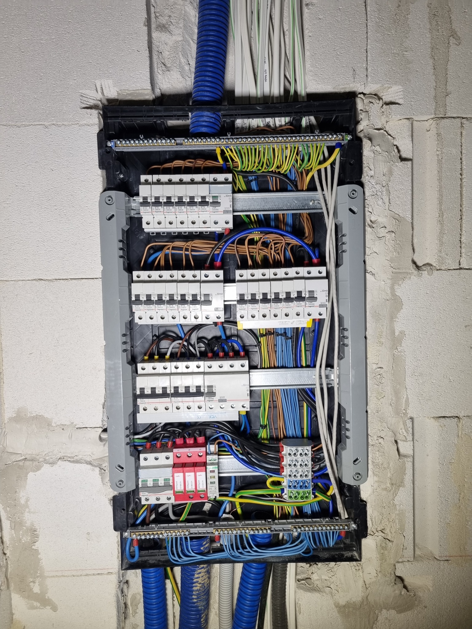Rozdzielnica elektryczna z zainstalowanymi bezpiecznikami, przewodami w kolorowych izolacjach i niebieskimi peszlami, zamontowana w surowej ścianie z betonu komórkowego.