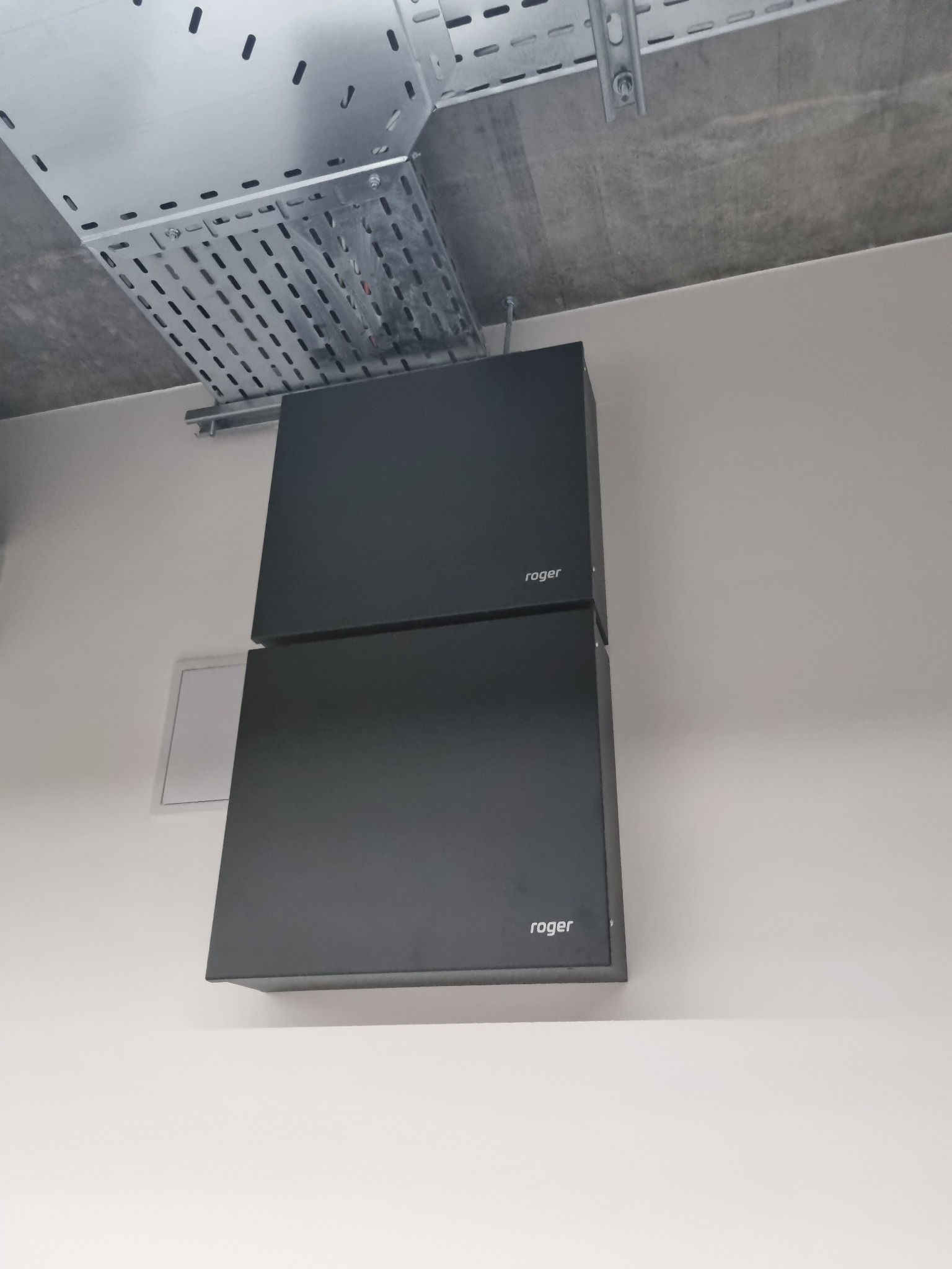 Dwa czarne moduły Roger zamontowane jeden nad drugim na jasnej ścianie, widoczna perforowana metalowa koryta kablowa pod sufitem.