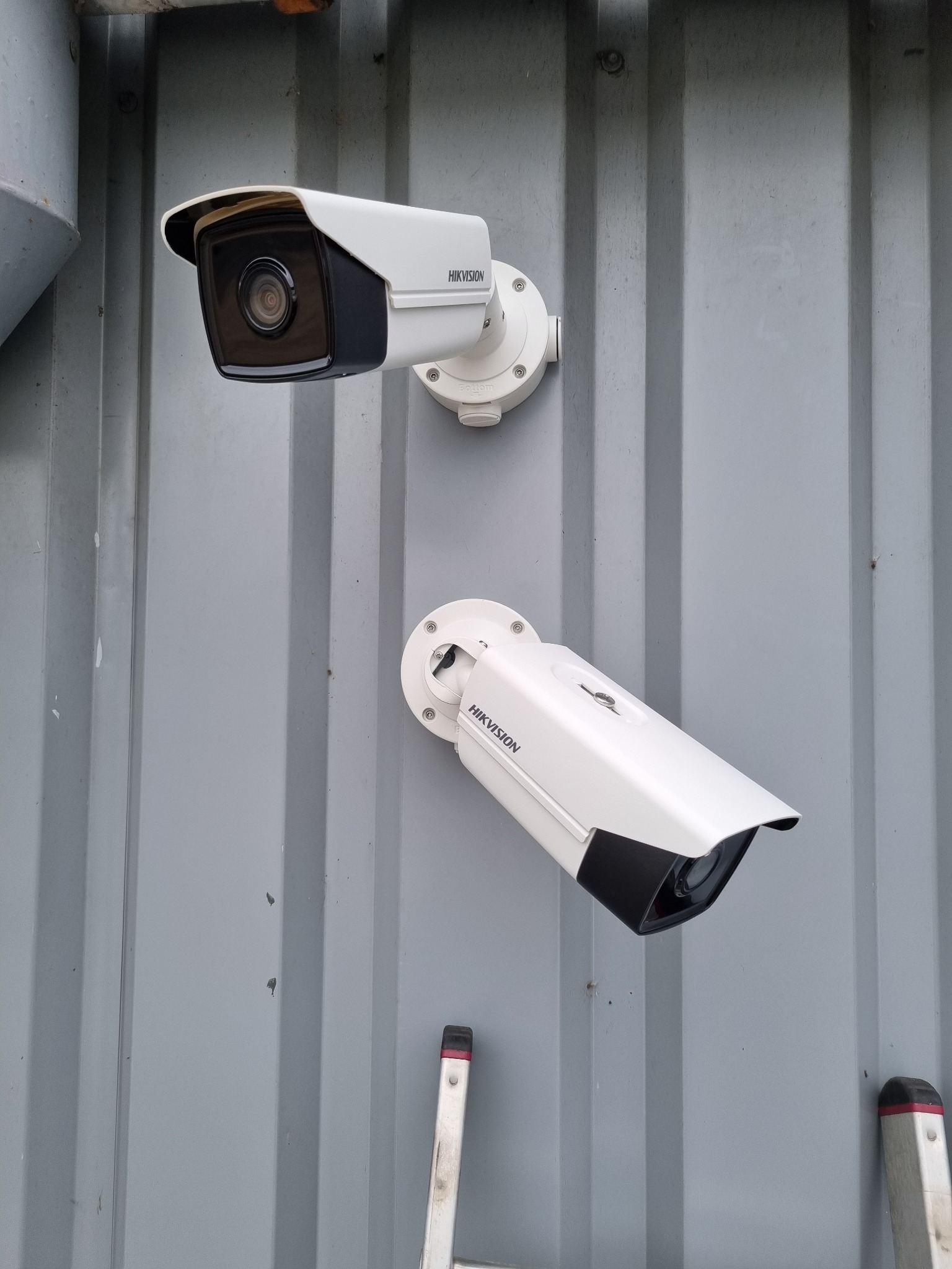 Dwie białe kamery przemysłowe marki Hikvision zamontowane na szarej, pionowo żłobkowanej ścianie kontenera, widoczny fragment drabiny w dolnym rogu.