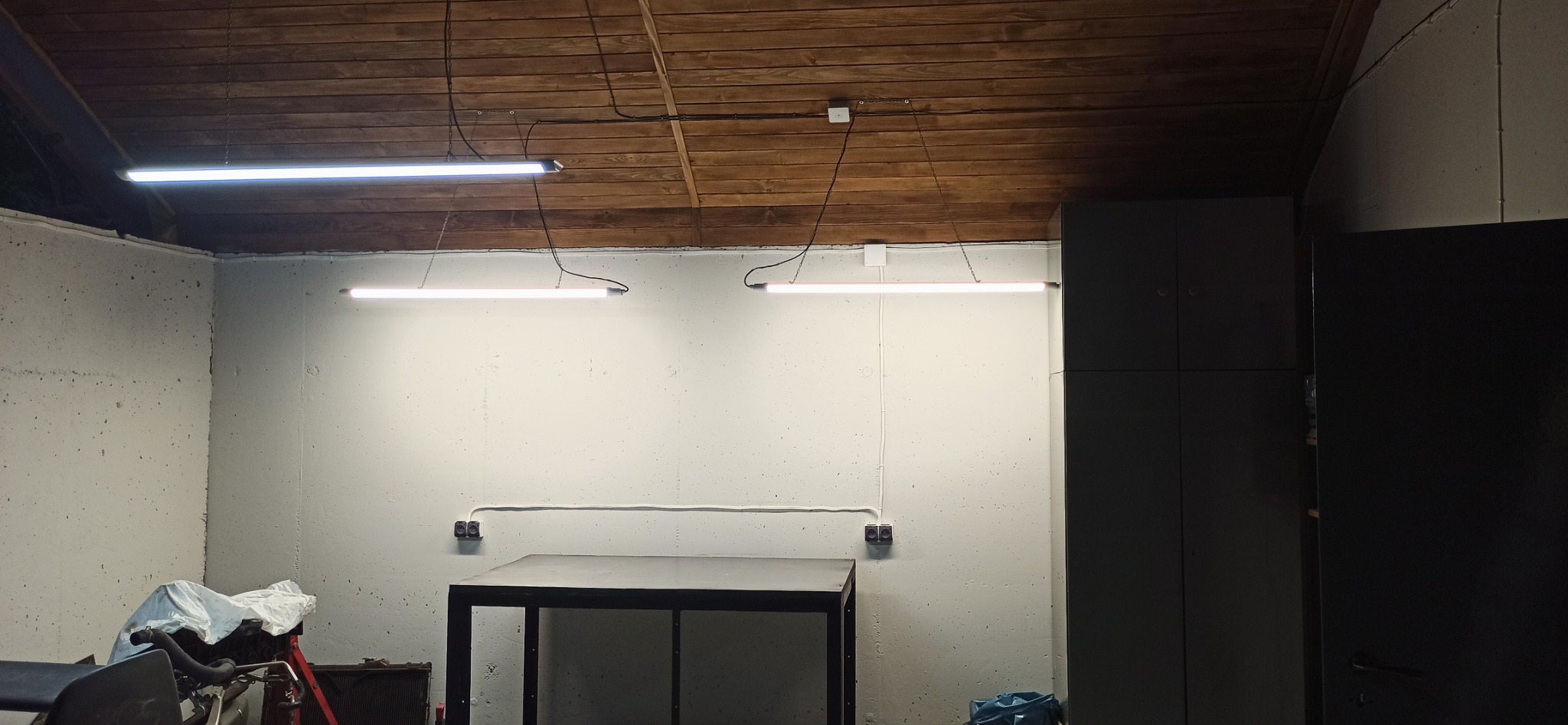 Wnętrze garażu z drewnianym sufitem, oświetlone dwiema liniowymi lampami LED zawieszonymi na łańcuchach, widoczny stół warsztatowy i szafa w tle.