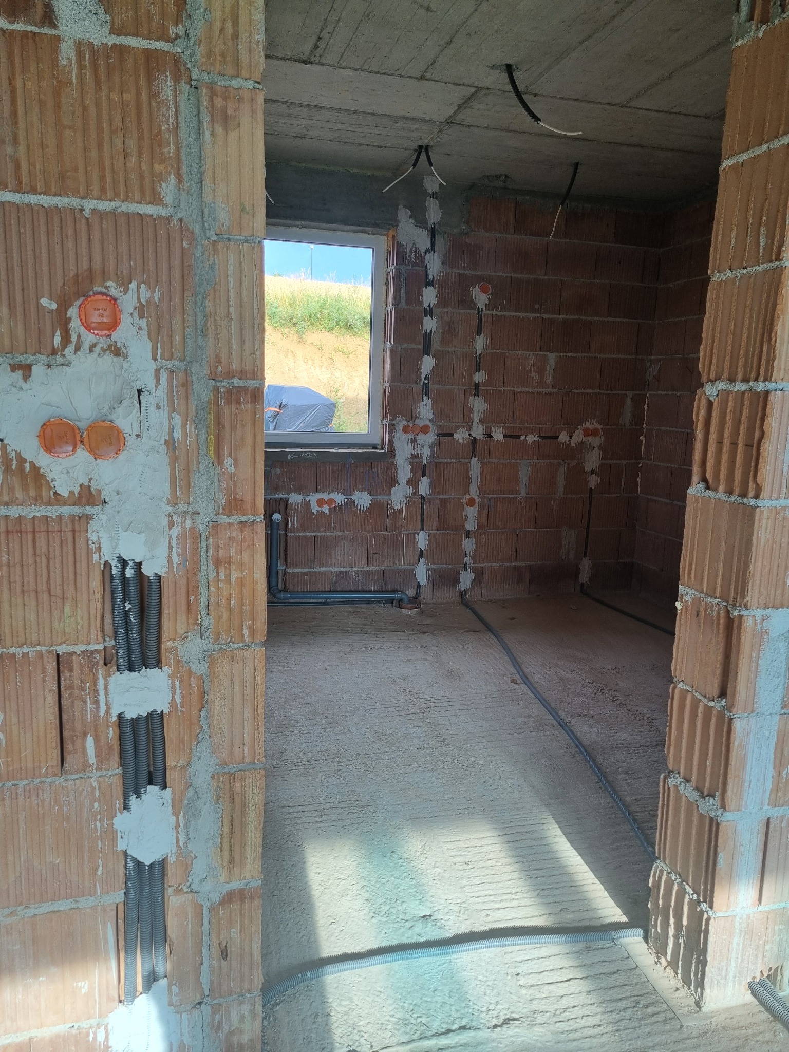 Surowy stan wnętrza budynku w trakcie prac instalacyjnych: widoczne przewody elektryczne w peszlach, puszki instalacyjne w ścianach z cegły, surowy betonowy sufit z wystającymi kablami, w tle widok...