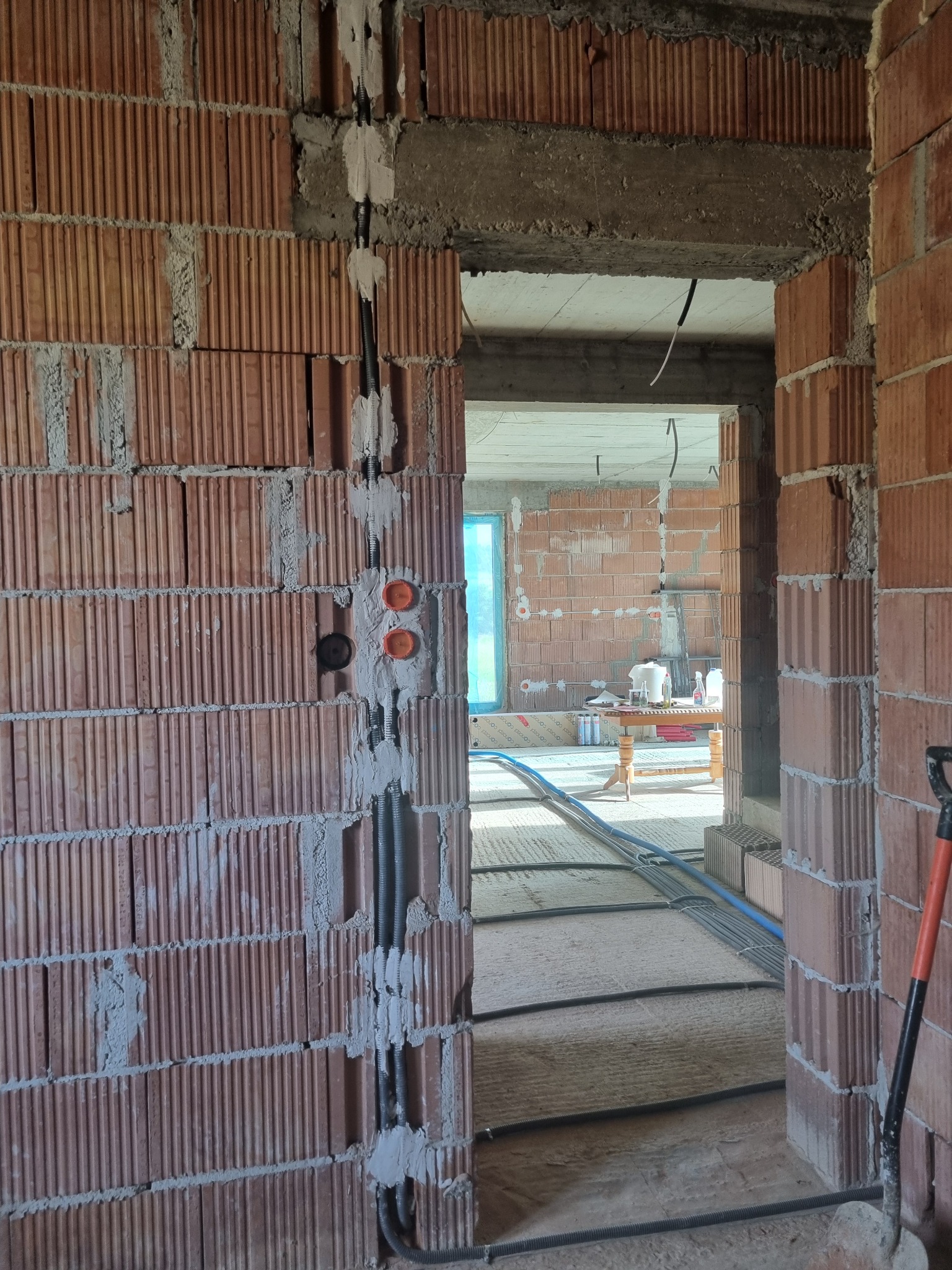 Surowy mur z czerwonej cegły z bruzdami wypełnionymi zaprawą, w których poprowadzone są czarne rury instalacyjne i puszki elektryczne, widok przez otwór drzwiowy na kolejne pomieszczenie w stanie...