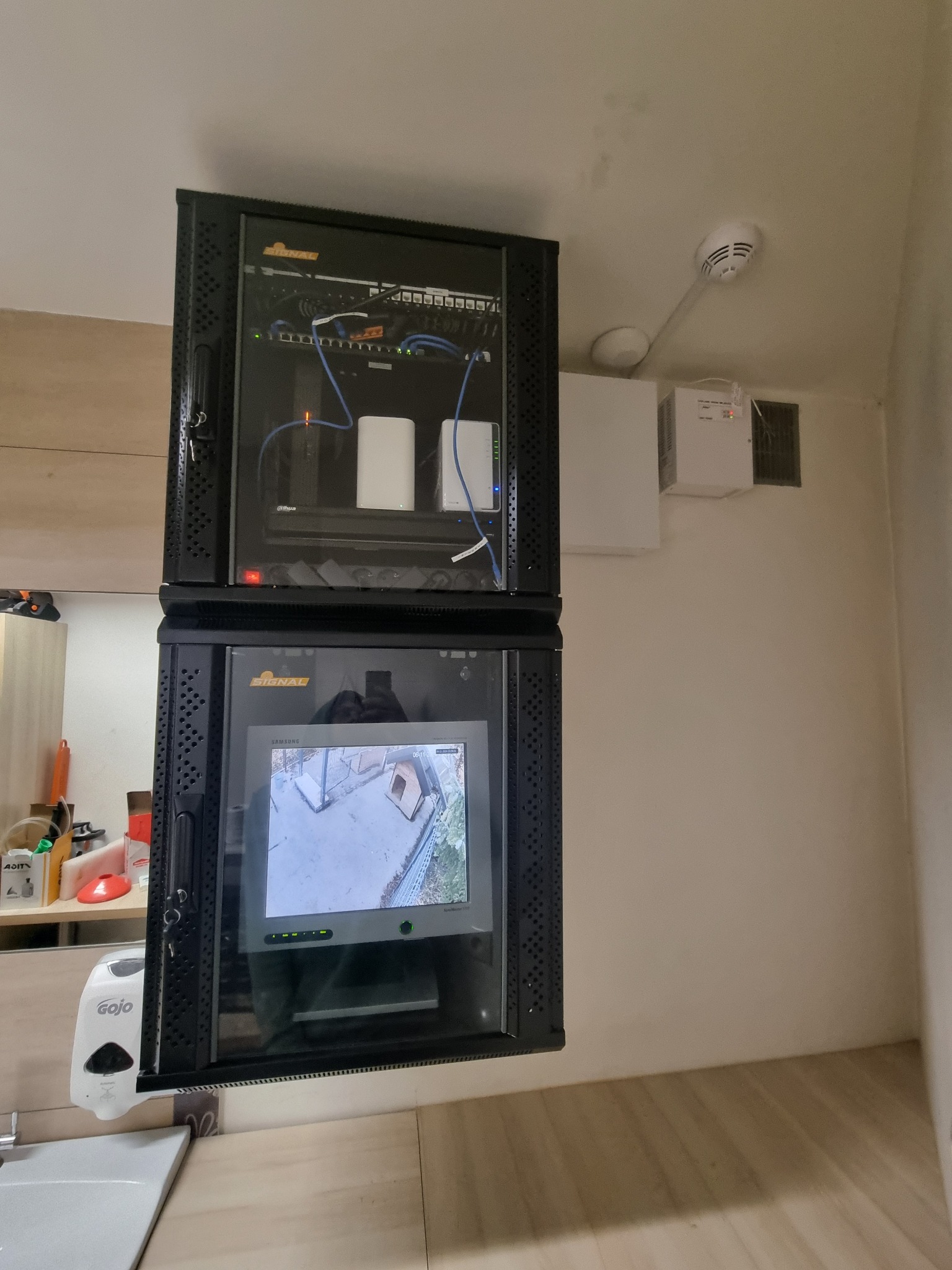 Dwa czarne, wiszące panele rack z zamontowanym sprzętem: górny z elementami sieciowymi i dolny z monitorem podglądu z kamery, obok czujnik dymu i biała skrzynka elektryczna na ścianie.