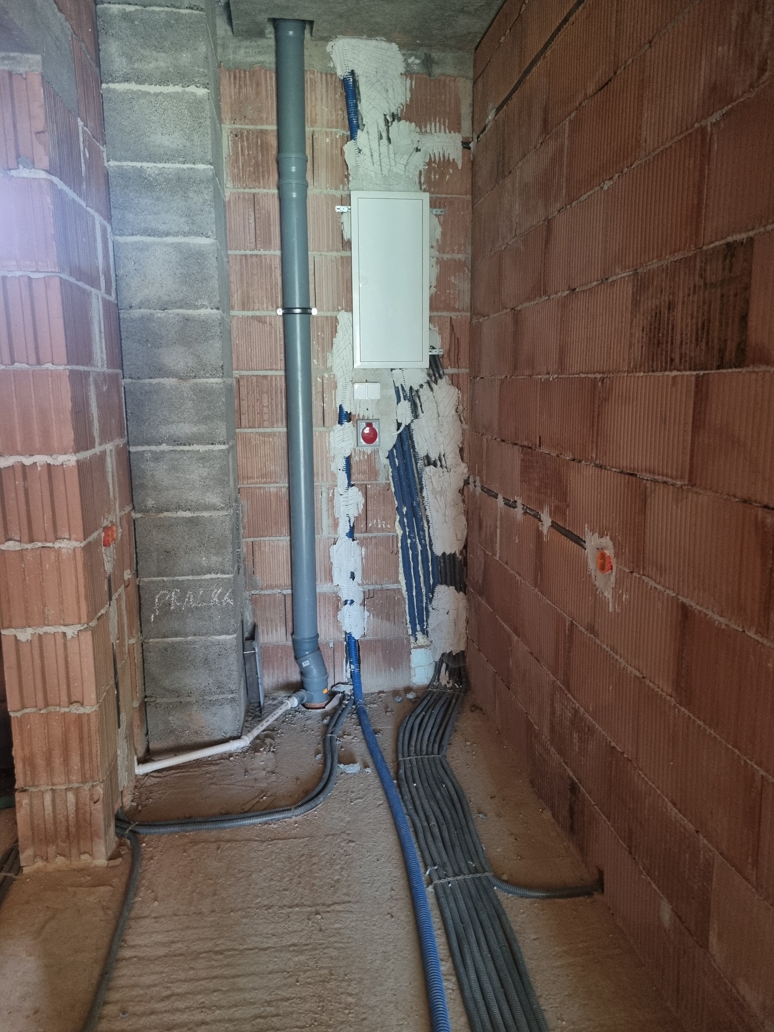 Surowy stan instalacji elektrycznej w budynku w trakcie budowy: widoczne kable w peszlach, puszka elektryczna, rury kanalizacyjne i ściany z czerwonej cegły dziurawki, na ścianie napis 'PRALKA'.