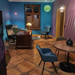 Biuro z drewnianym biurkiem, krzesłami i parkietem w geometryczny wzór. Na ścianach niebieski i fioletowy kolor. Okno z żaluzjami i lampy wiszące.