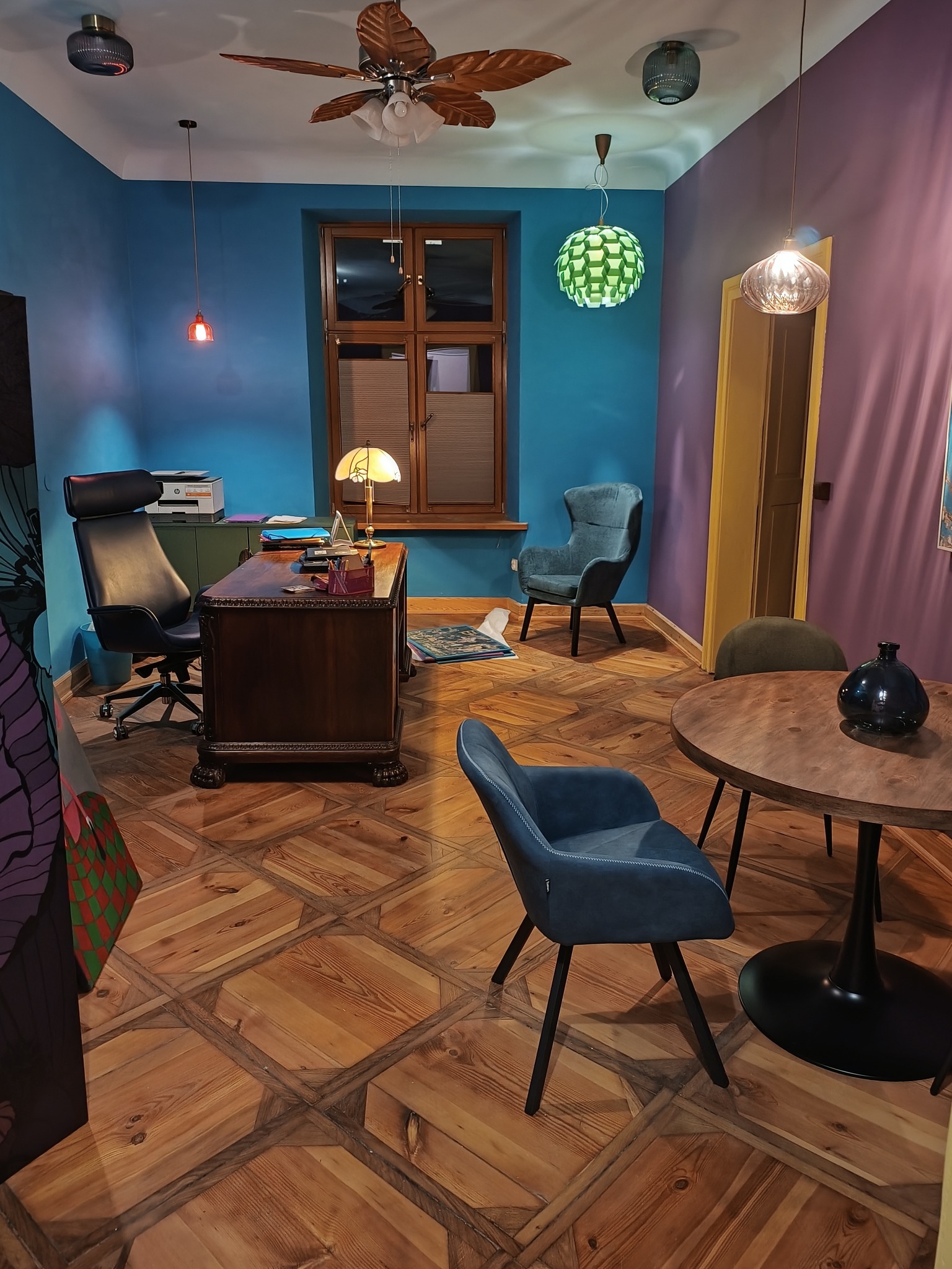 Biuro z drewnianym biurkiem, krzesłami i parkietem w geometryczny wzór. Na ścianach niebieski i fioletowy kolor. Okno z żaluzjami i lampy wiszące.