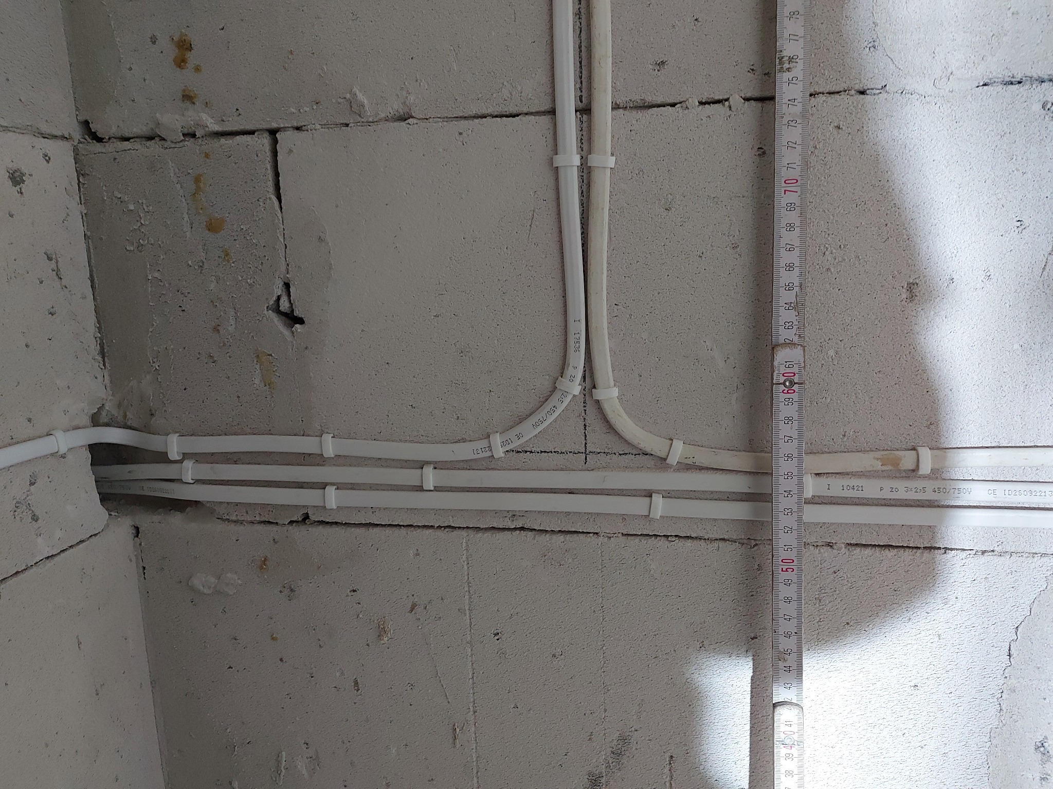 Białe rurki instalacyjne przymocowane do surowej ściany z betonu komórkowego, z widoczną miarką zwijaną od dołu.