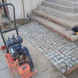 Wibrująca zagęszczarka obok fragmentu ułożonej szarej kostki granitowej przed betonowymi schodami, widoczne narzędzia: poziomica, miarka, młotek i szczotka.