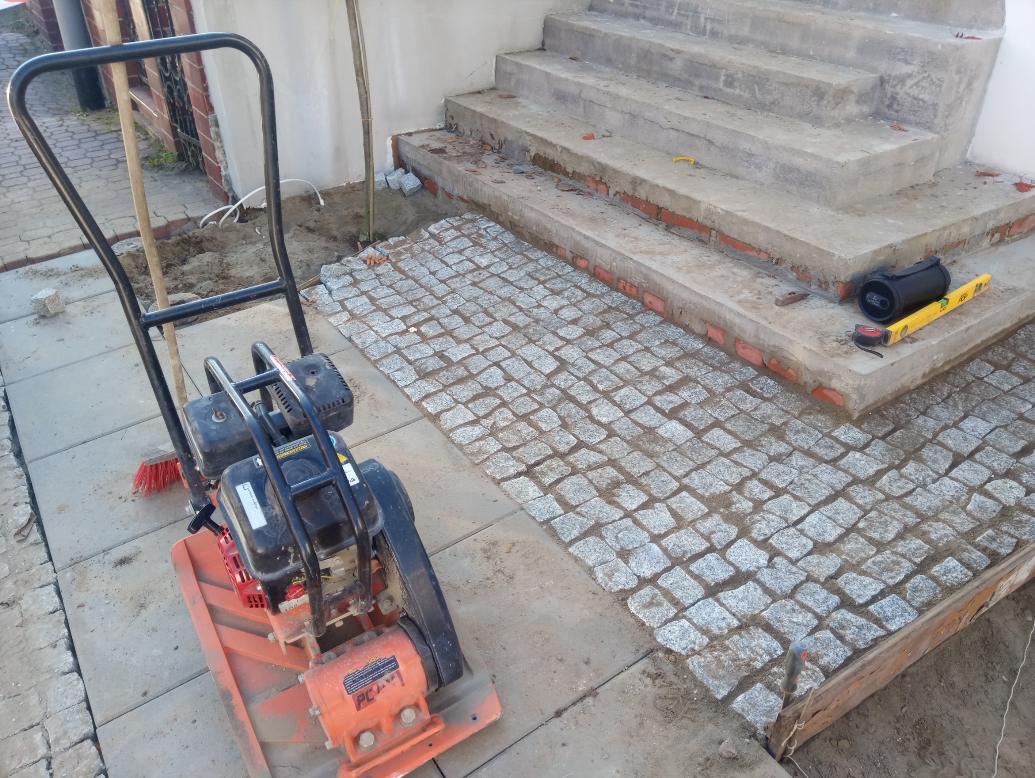 Wibrująca zagęszczarka obok fragmentu ułożonej szarej kostki granitowej przed betonowymi schodami, widoczne narzędzia: poziomica, miarka, młotek i szczotka.