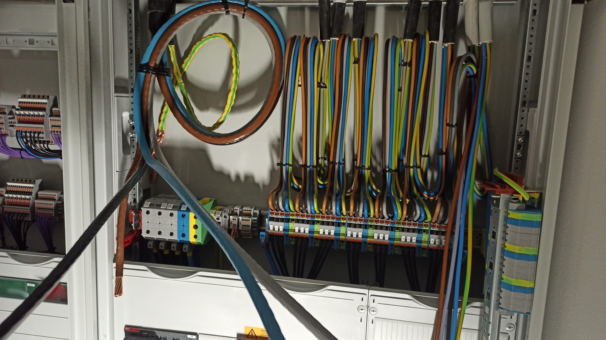 Rozdzielnica elektryczna z uporządkowanymi kolorowymi przewodami, widoczne złącza i zabezpieczenia, przewody ułożone w pętle i mocowane opaskami zaciskowymi, nowa instalacja.