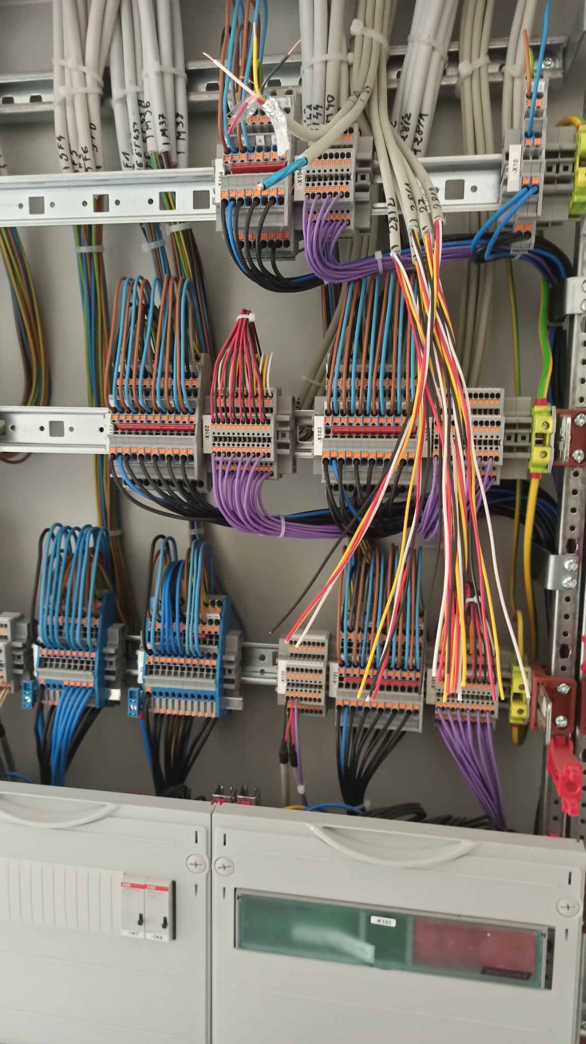 Szczegółowe ujęcie rozdzielnicy elektrycznej z widocznymi przewodami o różnych kolorach, podłączonymi do listew zaciskowych, z opisami na przewodach i listwach, wewnątrz szarej obudowy.