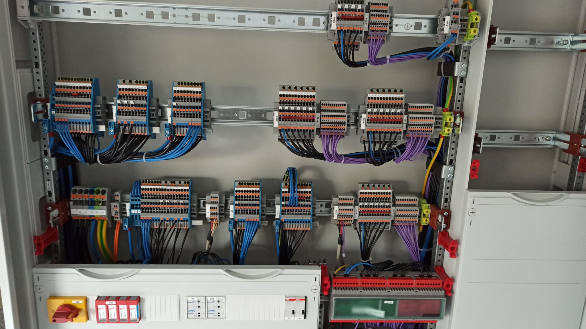 Rozdzielnica elektryczna z licznymi złączkami, przewodami w różnych kolorach (niebieski, czarny, fioletowy, żółty, zielony, pomarańczowy) oraz zabezpieczeniami, zamontowana w metalowej obudowie...