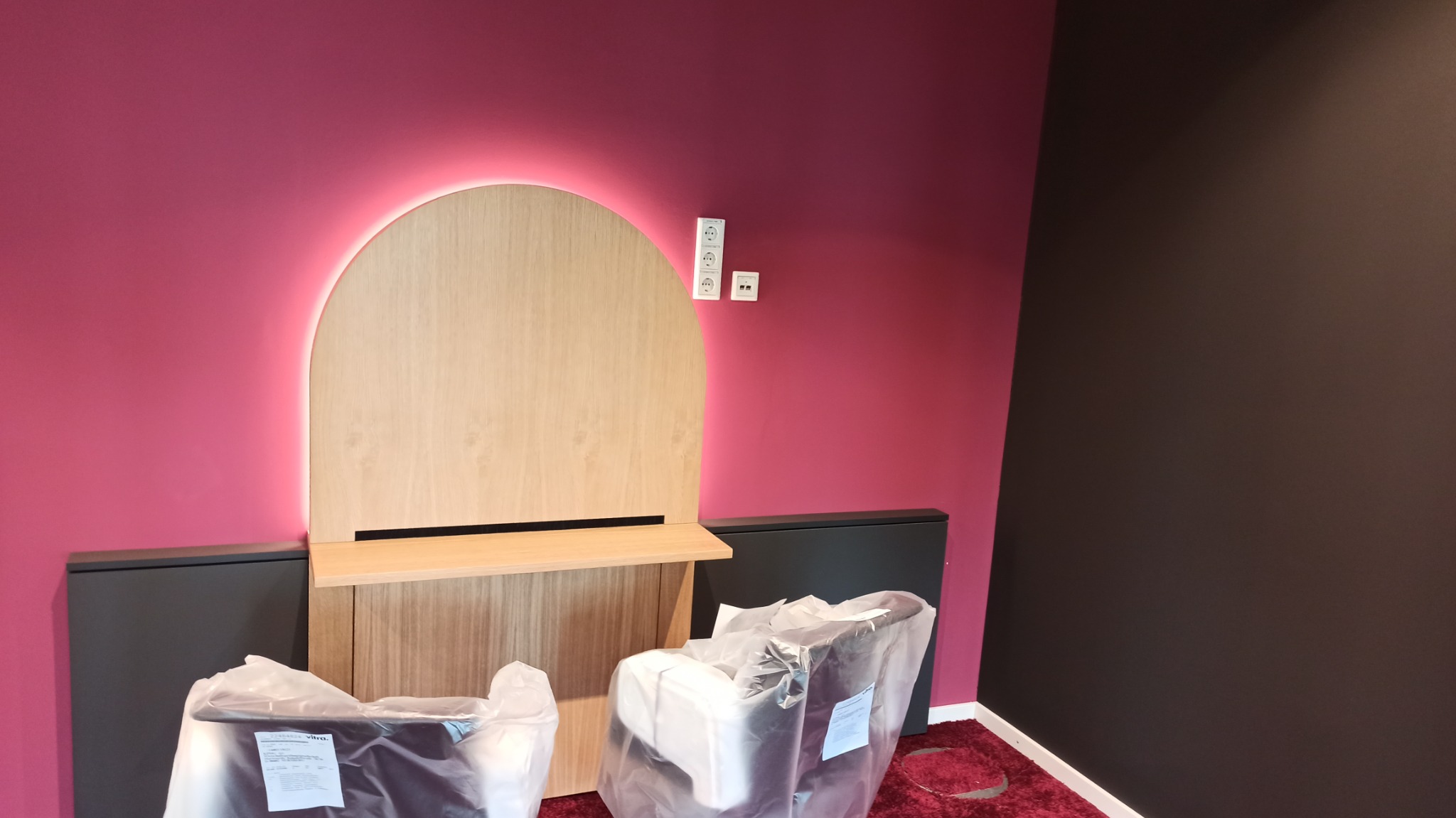 Nowoczesny pokój z bordową ścianą, podświetlanym drewnianym panelem z półką i gniazdkami elektrycznymi, z krzesłami zabezpieczonymi folią ochronną.