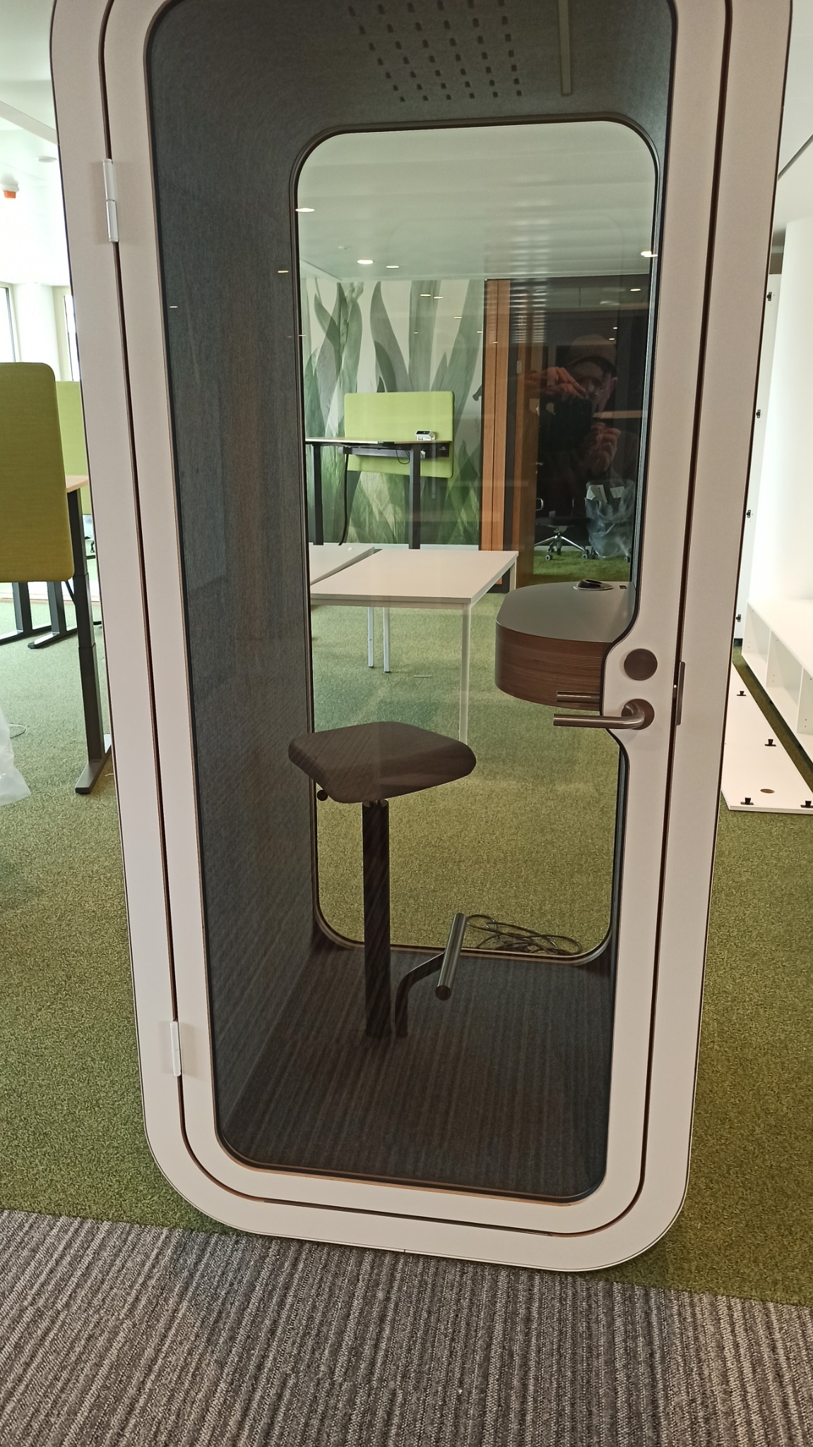 Nowoczesna, dźwiękoszczelna kabina biurowa z szarą tapicerką wewnętrzną i obrotowym krzesłem, widoczna na tle biura z zielonymi akcentami, odzwierciedlona w szklanej powierzchni kabiny.