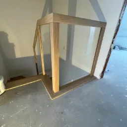 Szklana balustrada na schodach z drewnianymi elementami konstrukcyjnymi, widok od dołu, w tle fragment schodów i ściana w trakcie remontu.