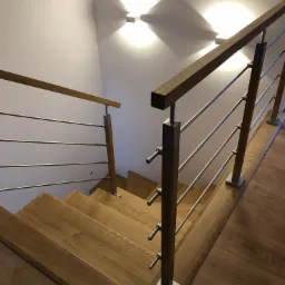 Jasne, drewniane stopnie schodów z nowoczesną balustradą ze stali nierdzewnej i drewnianą poręczą, oświetlone przez dwa kwadratowe plafony na białej ścianie.