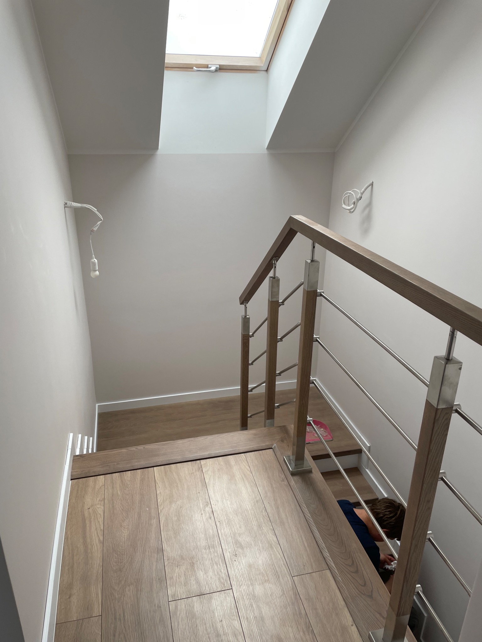 Wnętrze klatki schodowej z drewnianymi stopniami i poręczą z elementami stali nierdzewnej, widok z góry, w tle okno dachowe i wystający kabel z żarówką.