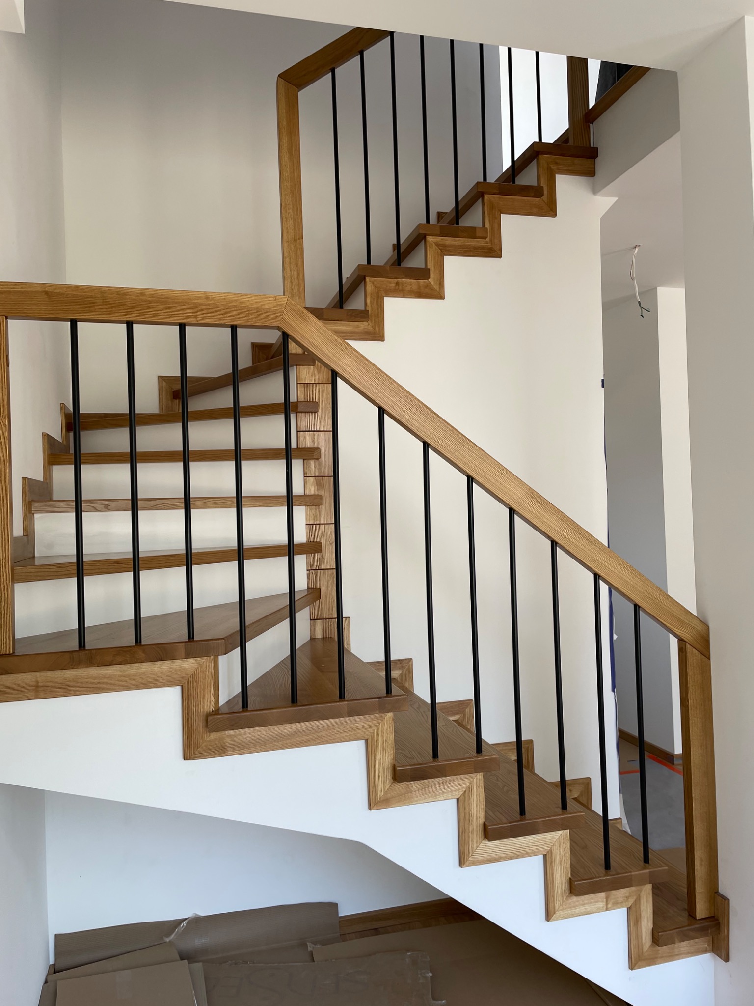 Drewniane schody z jasnego drewna z czarnymi, pionowymi balustradami, widok z dołu, częściowo zakryte kartonami na dole.