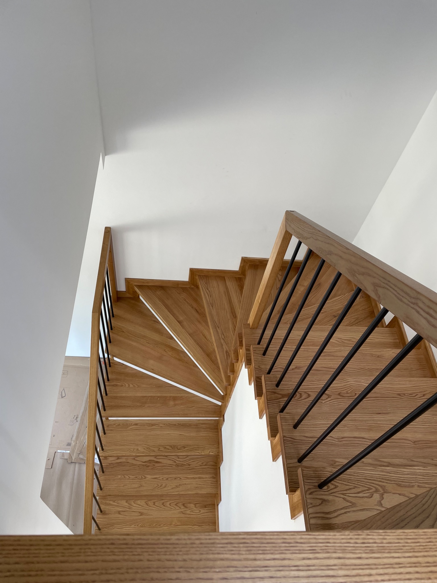 Widok z góry na spiralne schody z jasnego drewna z czarnymi metalowymi poręczami, w minimalistycznym wnętrzu z białymi ścianami.