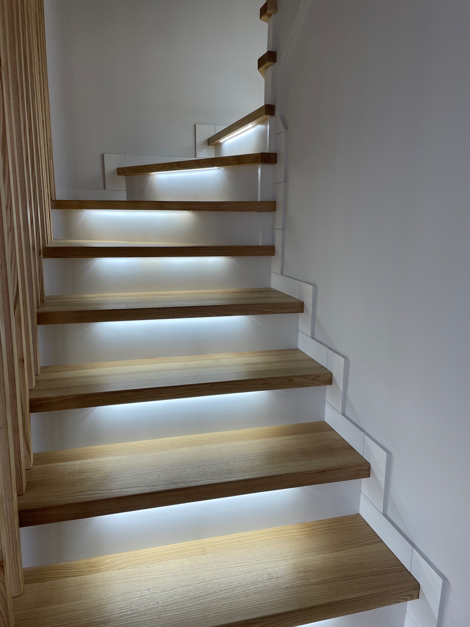 Nowoczesne schody z jasnego drewna z podświetleniem LED pod każdym stopniem, minimalistyczna biała ściana i drewniana balustrada z pionowych listew.