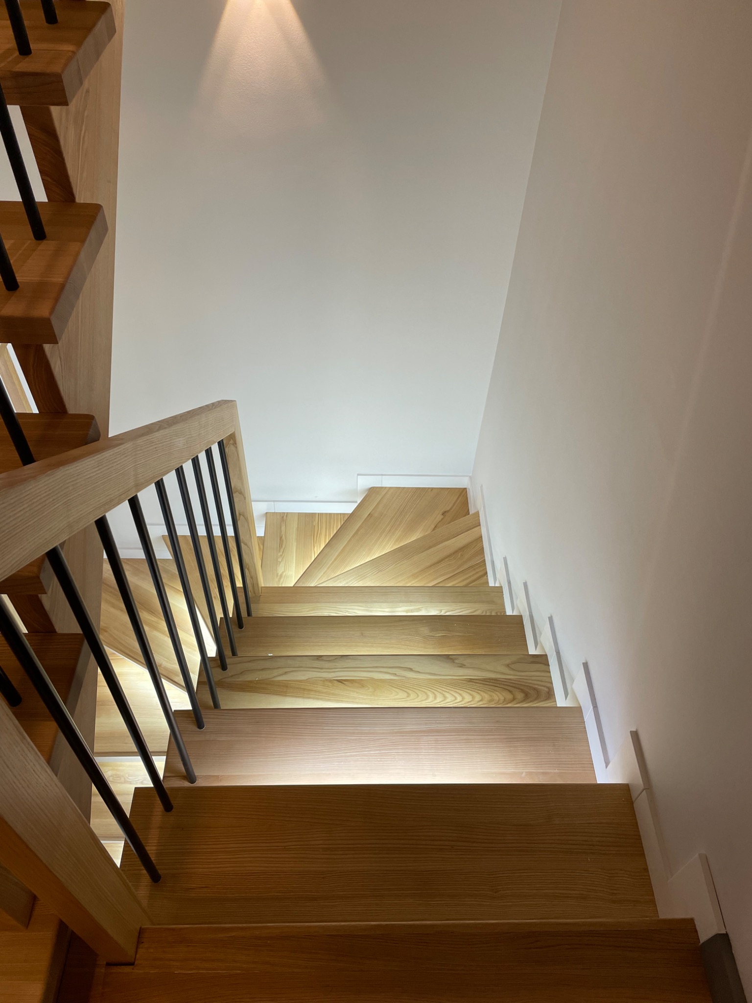 Widok z góry na nowoczesne, jasne schody z drewna z czarnymi, metalowymi balustradami i subtelnym oświetleniem LED pod stopniami, tworzącymi ciepły blask na białej ścianie.