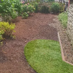 Fragment ogrodu z wyściółkowaną korą rabatą kwiatową, ułożoną trawą z rolki i obrzeżem z kamyków przy ścianie budynku.